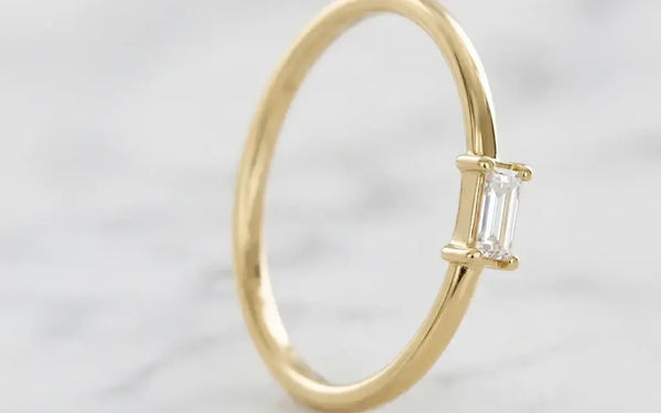 Solitaire baguette diamond engagement ring in 18K yellow gold by Mayuri, on an elegant background