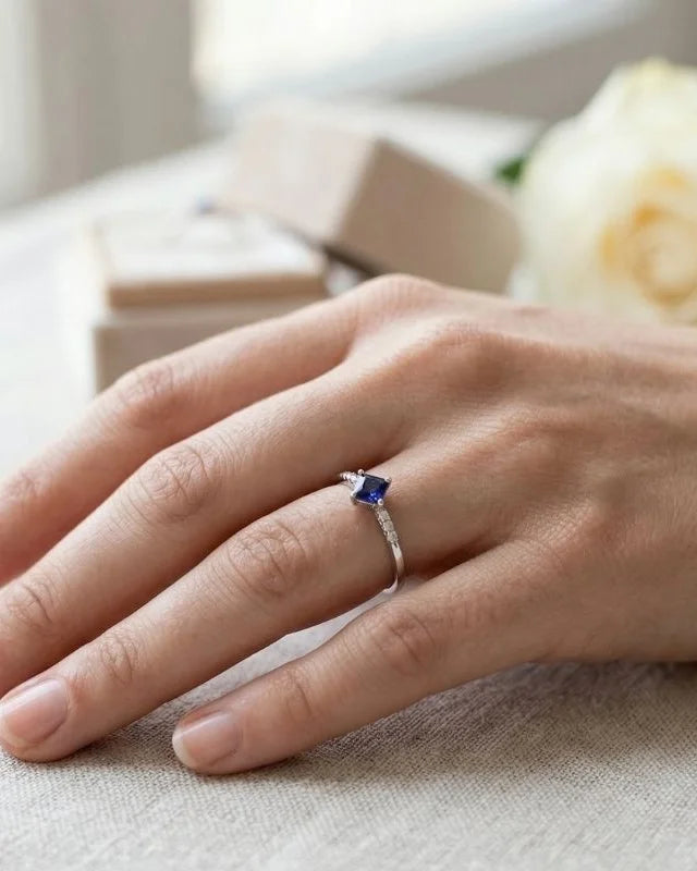 18K white gold ring set with a princess cut blue sapphire and diamonds, worn on the ring finger