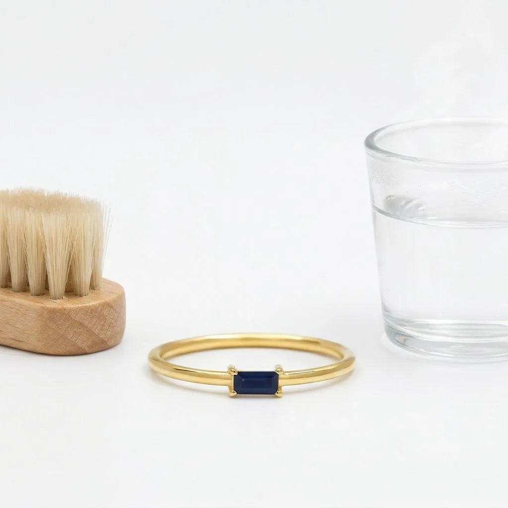 Jewellery cleaning materials featuring a sapphire ring, a soft brush, and a glass of water on a neutral background