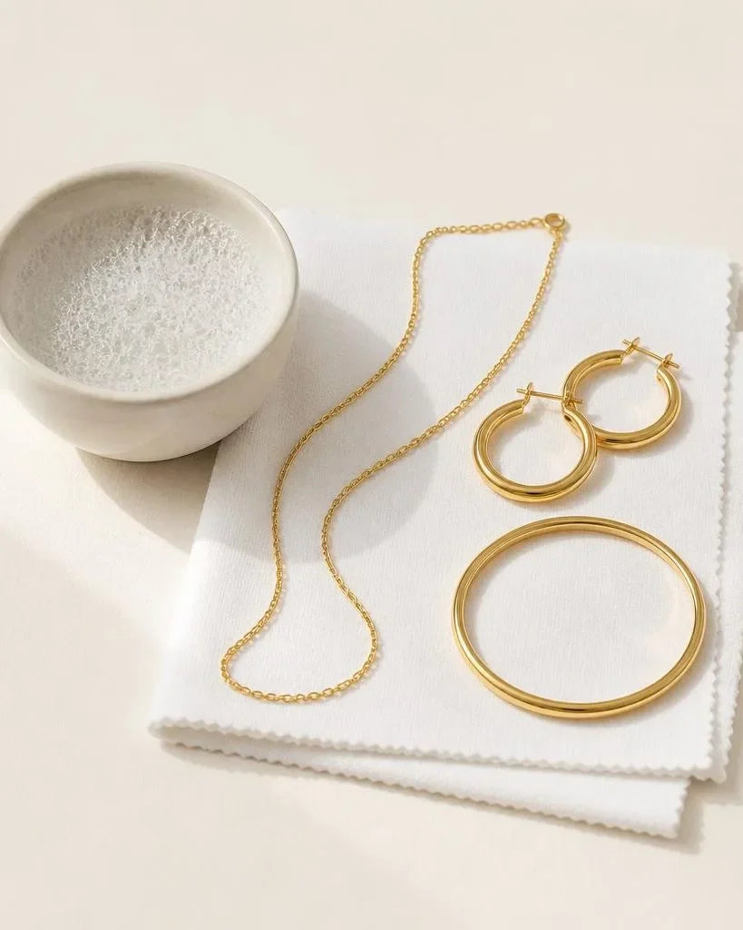 Gold-plated jewellery arranged on a microfibre cloth beside a bowl of soapy water, ready for gentle cleaning
